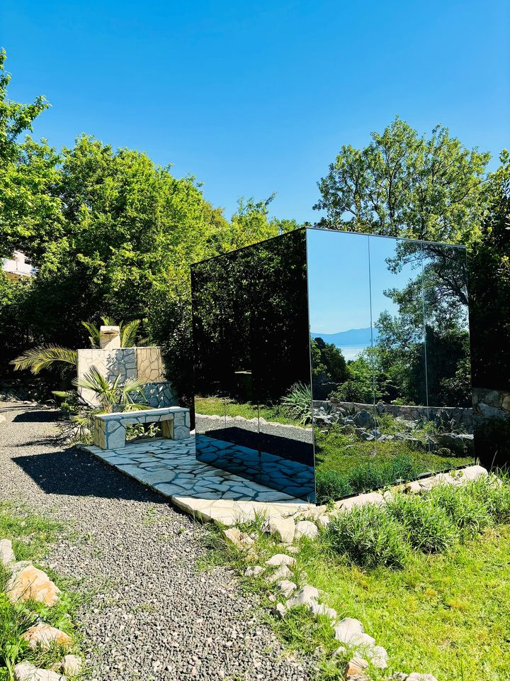 The mirrored sauna cube in daylight, reflecting the green canopy and a sliver of the Adriatic